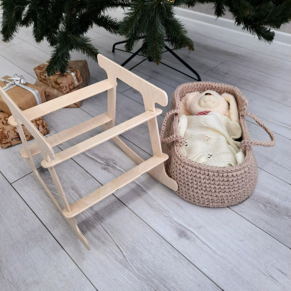 In a beige handmade basket for dolls lies a toy hare, which is covered with a milky-white knitted blanket, and next to it stands a wooden stand for this basket