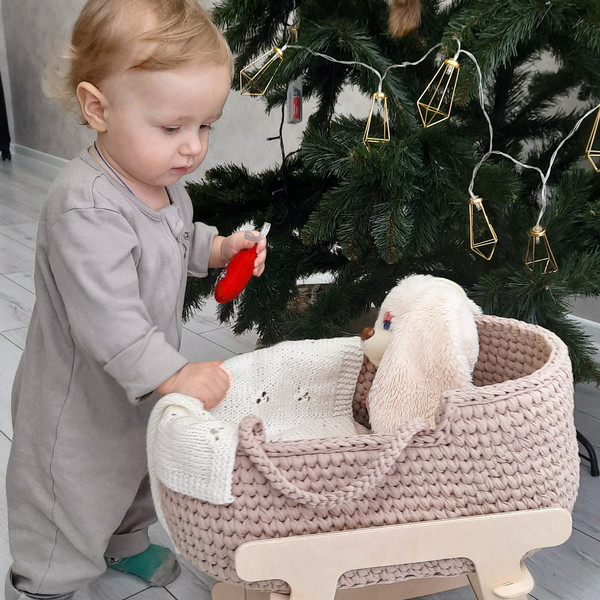 A one-year-old girl rocks a baby doll basket in beige with a milky-white hand-knitted blanket on a wooden stand